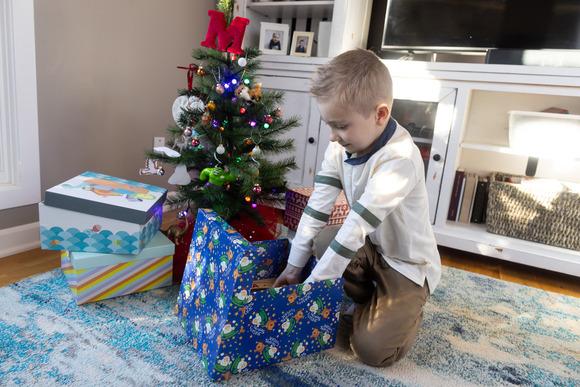 Boy in a living room in front of a Christmas tree opening a wrapped box 