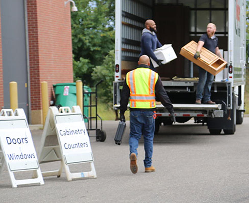 Three men loading cabinets into box truck next to signs saying doors, windows, cabinetry, counters