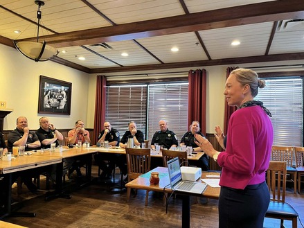 Cmsr. Edelson speaks to a group of police and fire chiefs sitting at a table in a "C" position in a large dining room; they are listening.