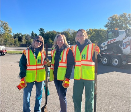 Heather and her staff smile in a photo while wearing neon vests and hold tree planting tools. They're on the side of the road.