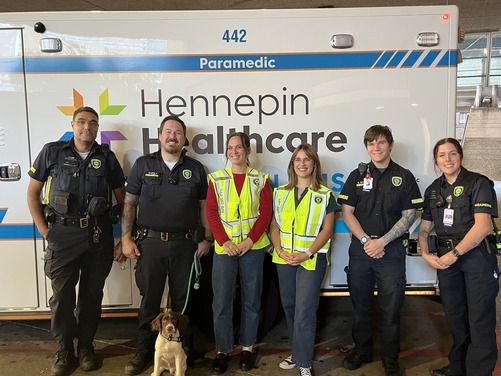 Heather's staff, Raina and Clara, stand smiling in front of an ambulance with an EMS emotional support dog and several paramedics.