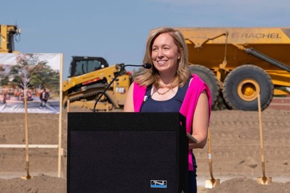 Heather speaks at a podium, wearing a pink scarf, in front of a construction site at the Southdale Library site.
