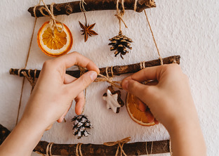 Hands are shown assembling sustainable holiday decor with strings, sticks, dried oranges, and pinecones.