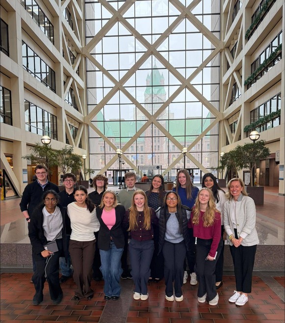 Heather poses with 14 students, smiling, in two rows in the Gov. Center lobby. They are all smiling.