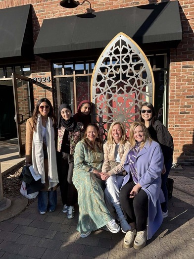 Heather sits for a photo, smiling, with six other women. They sit on the sidewalk at 50th and France and it is sunny.