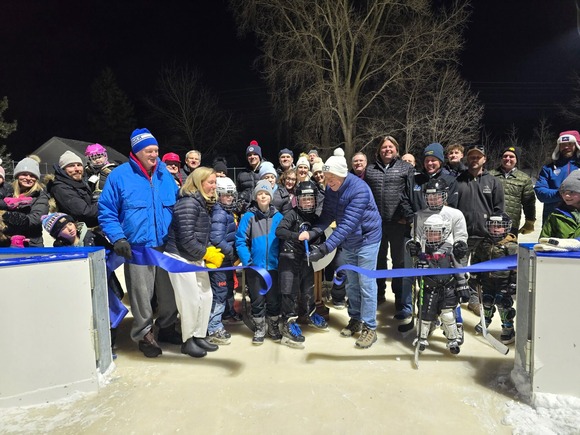 Heather claps, smiling, and is surrounded by Deephaven mayor Kent Carlson and many young hockey players as the mayor cuts the ribbon.