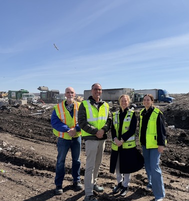 Commissioner Edelson and Commissioner Anderson stand at the landfill with trucks in the distance and two staff, all in neon vests.