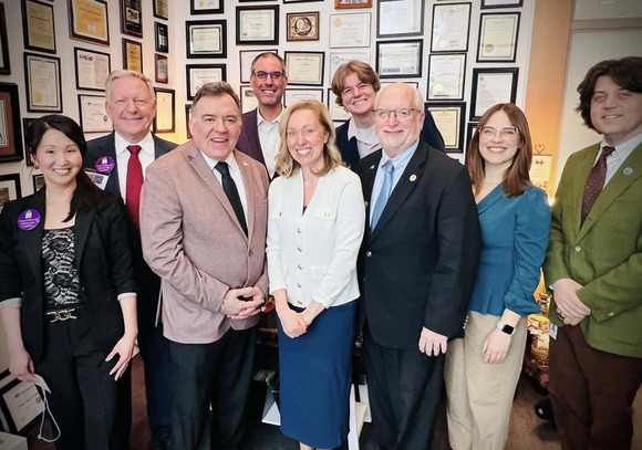 Cmsr. Edelson poses with Cmsr. Anderson and staff, smiling, with Senator John Hoffman in his office. The background is a couch and many photos.