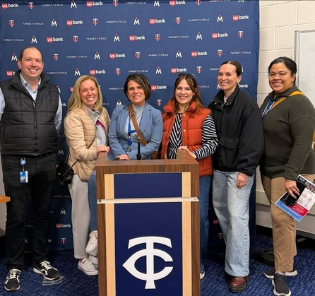 Cmsr. Edelson poses in front of a Twins-branded podium with staff and Administrator Wentland, all smiling. 6 people around the podium.