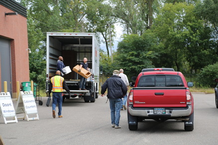 A man drops off materials from the back of a red truck at a pick-up event outside of a building.