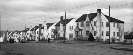 Two-story white houses lining a street with stairs leading from sidewalk, cars parked out front, and a person on sidewalk