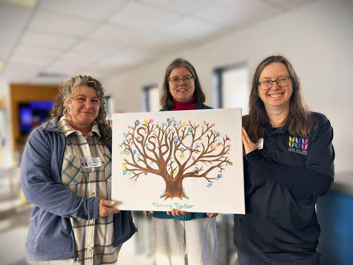 Three women holding a painting of a tree with multi colored thumbprints