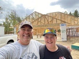 Two people wearing hats and T-shirts stand with their arms around each other in front of a construction frameup.