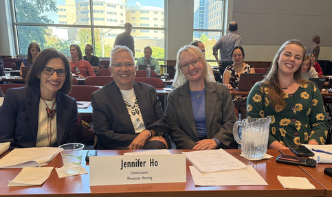 Four people sitting at a desk, including Lt. Governor Peggy Flanagan and Commissioner Jennifer Ho. Behind them are tables, people and tall windows.