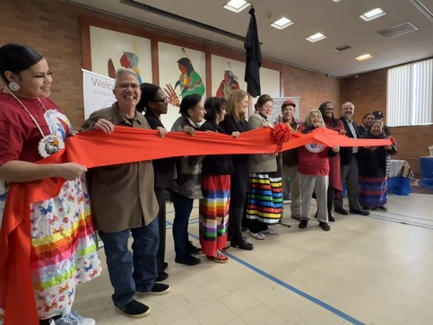 A dozen people stand in a line holding a red ribbon in a room with painted panels of Native women in the background.