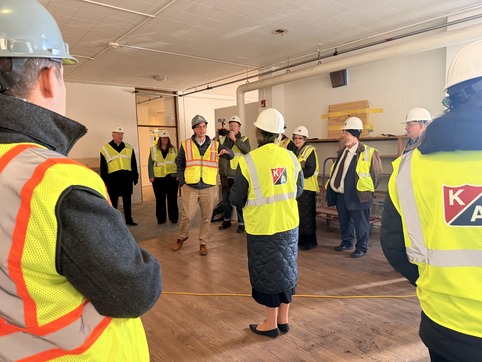 Group of people in hard hats and reflective vests listening to a speaker during a site visit indoors.