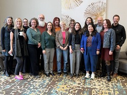 Fifteen people in business casual pose for a group photo on multicolored carpeting against a wall.