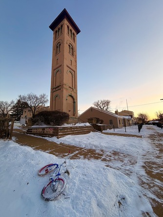 A kids' bike leans on a snowbank in front of a tall tower on a bright, cold day.