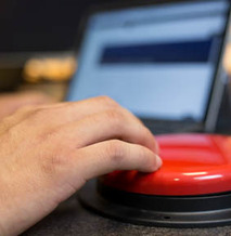 Hand pressing an assistive technology switch in front of a computer.