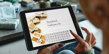 Woman typing typing text onto a digital holiday card on a tablet, presents in the background.