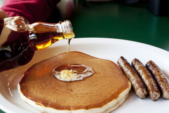 A golden brown pancake with maple syrup being poured on top. There are sausage links on the side.