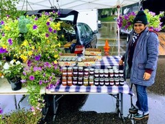 A woman stands with her farmers market table selling flowers and preserves.