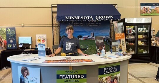 A woman in a blue shirt stands at a booth representing Minnesota Grown at the state fair.