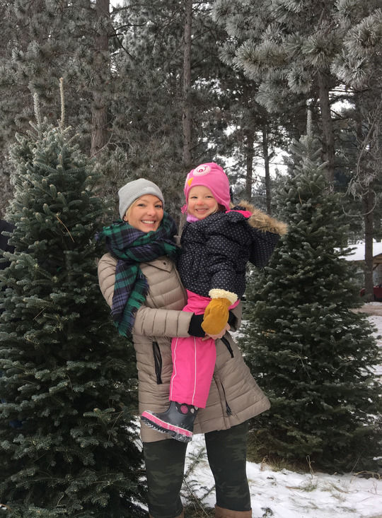 A woman holding a young girl standing out in a snowy field of Christmas trees.