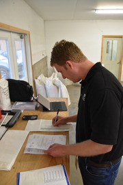 An inspector in a black shirt and jeans is standing at a table with pen in hand while reviewing a paper seed label. 