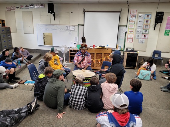 A man wearing American Indian accessories sits at a drum with nine boys as other students watch sitting arranged around them on the flor