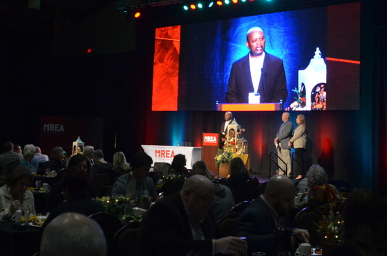 Commissioner Jett is seen speaking at a podium and on a large screen behind him