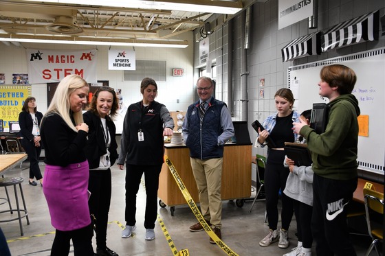 A teacher points out something students are doing behind a crime scene tape in the Criminalistics section of the STEM Lab