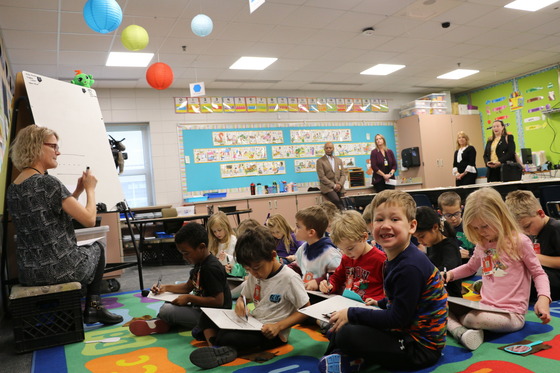 A boy smiles at the camera as his teacher teaches the lesson and Commissioner Jett, Julie Novak and other adults observe the lesson