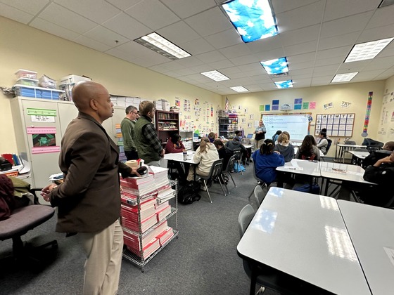 Commissioner Jett stands at the back of the room as a teacher teaches a math lesson at the front of the room