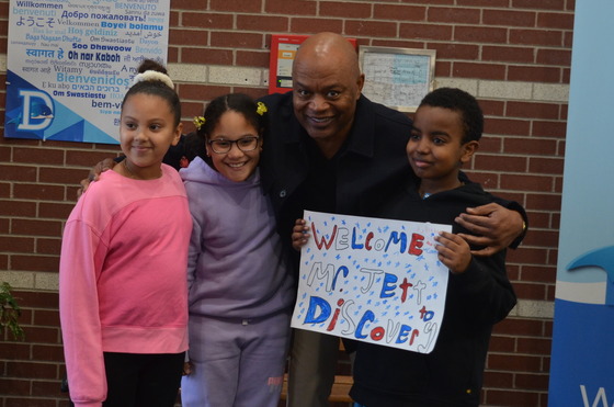 Commissioner Jett stands with three children who are holding a sign that says "Welcome Commissioner Jett" 