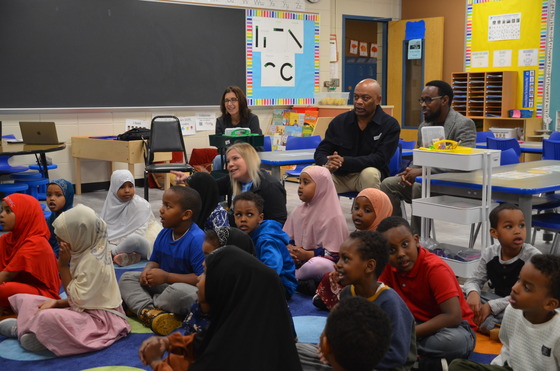 Commissioner Jett and two adults sit at the back of a classroom as students sit on the ground for a lesson