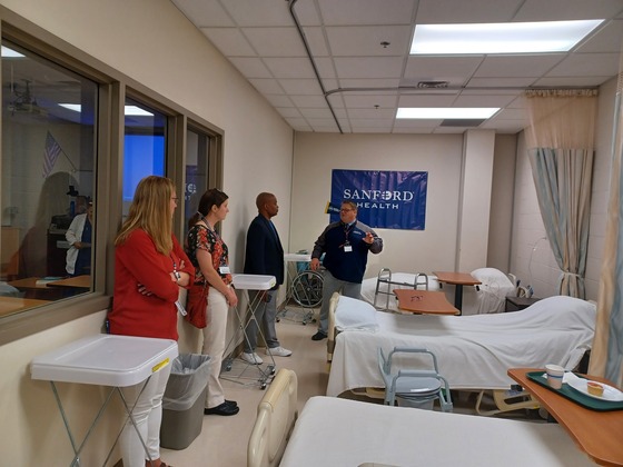 Commissioner Jett and others stand in a hospital style room with a sign that says Sanford Health hanging on the wall