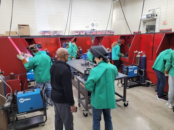 A student speaks to Commissioner Jett as they look on at several students doing welding in welding bays