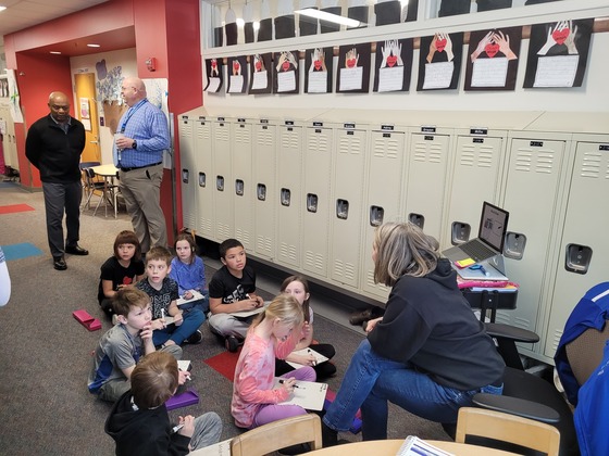 A teacher teaches a lesson to students seated on the ground as Commissioner Jett looks on 