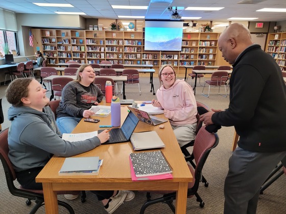 Three students siting at a table in a library look up at Commissioner Jett while he speaks to them