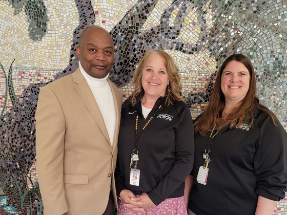 Commissioner Jett stands with two women from Nay Ah Shing Schools in front of a mural 