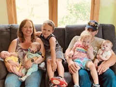 Nikki, a middle-aged woman with pale skin and red hair, sits on a couch with several children in front of a sunny window.