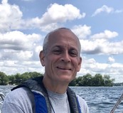 Image of a middle-aged man with short-cropped hair outside. He's wearing a life vest and a lake with a blue sky is in the background.