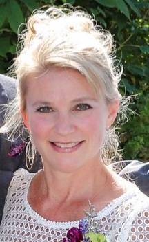 Shannon, a middle-aged with blonde hair pulled into an updo smiles at the camera. She's outside and wearing a white top with a flower boutineer.