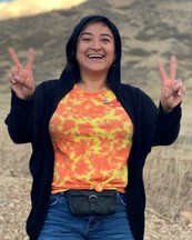 Rosa, a younger woman with dark hair and olive skin, holds her hands up in two peace signs outside.