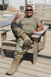 Fernando, a man with close-cropped dark hair and olive skin, smiles as he lounges in a wooden chair outdoor. He's wearing fatigues and sunglasses.