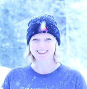 Daphne, a blonde woman with pale skin and blue eyes, stands in a snowy landscape. She's wearing a black beanie and a purple sweatshirt.