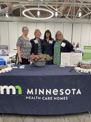 Image of 4 Caucasian women standing behind a table with navy colored MDH Health Care Homes table cover on it 