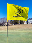 Yellow flag waving in breeze with logo Par 3 Community Golf Course over hole green near bare trees and bright blue sky. 