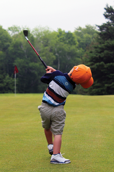 Boy with club in motion shown from back with orange golf cap, striped shirt and khaki shorts, swinging toward hole.